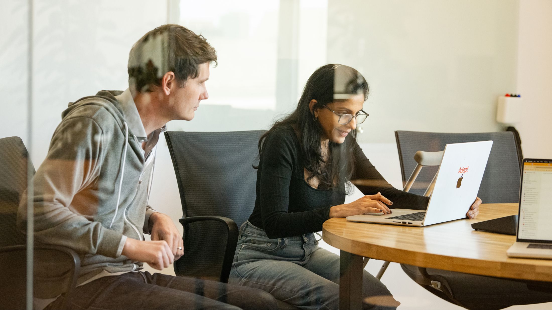 A photo of two team mates sat working together, one is working at a laptop.